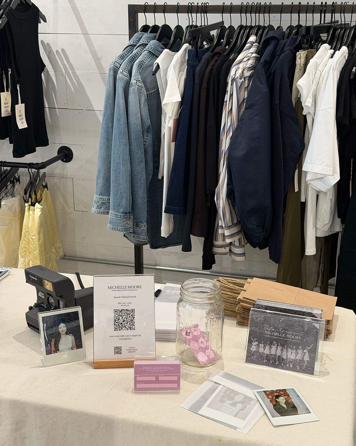 A clothing rack display with denim jackets and dark garments beside a table with Polaroid photos, a mason jar, and marketing materials.