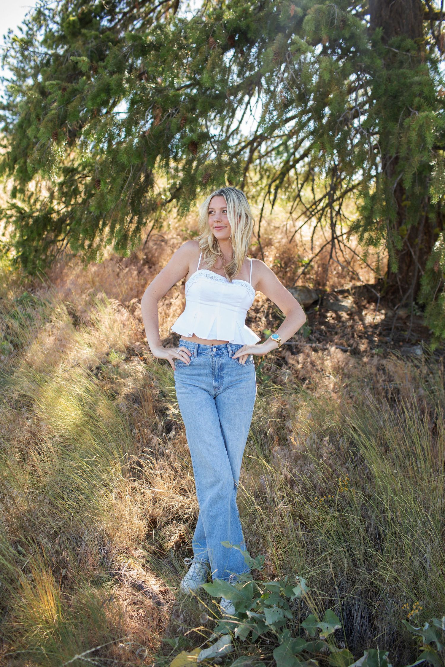 Blonde woman in white corset top and blue jeans posing with hands on hips among tall grass and trees.