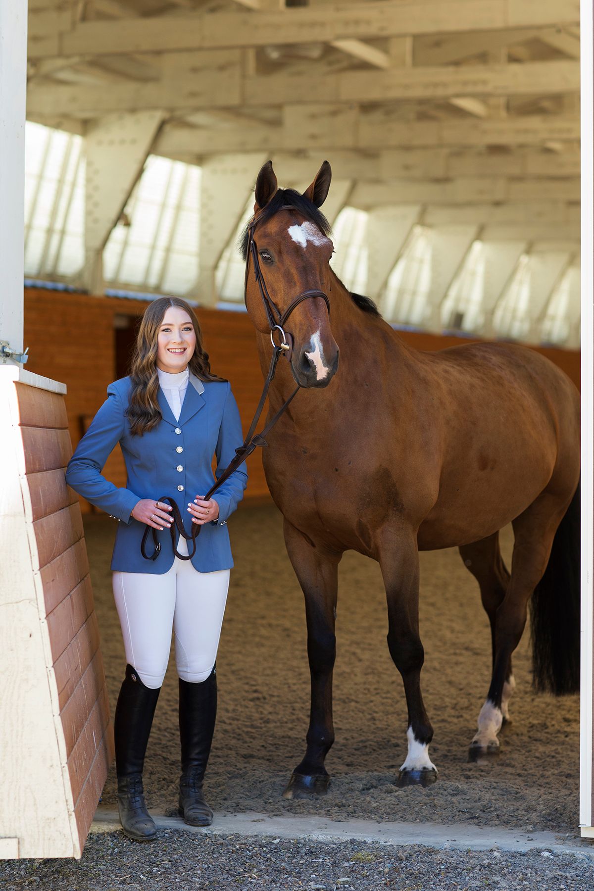 Young woman in blue equestrian jacket stands beside a chestnut horse at stable entrance.