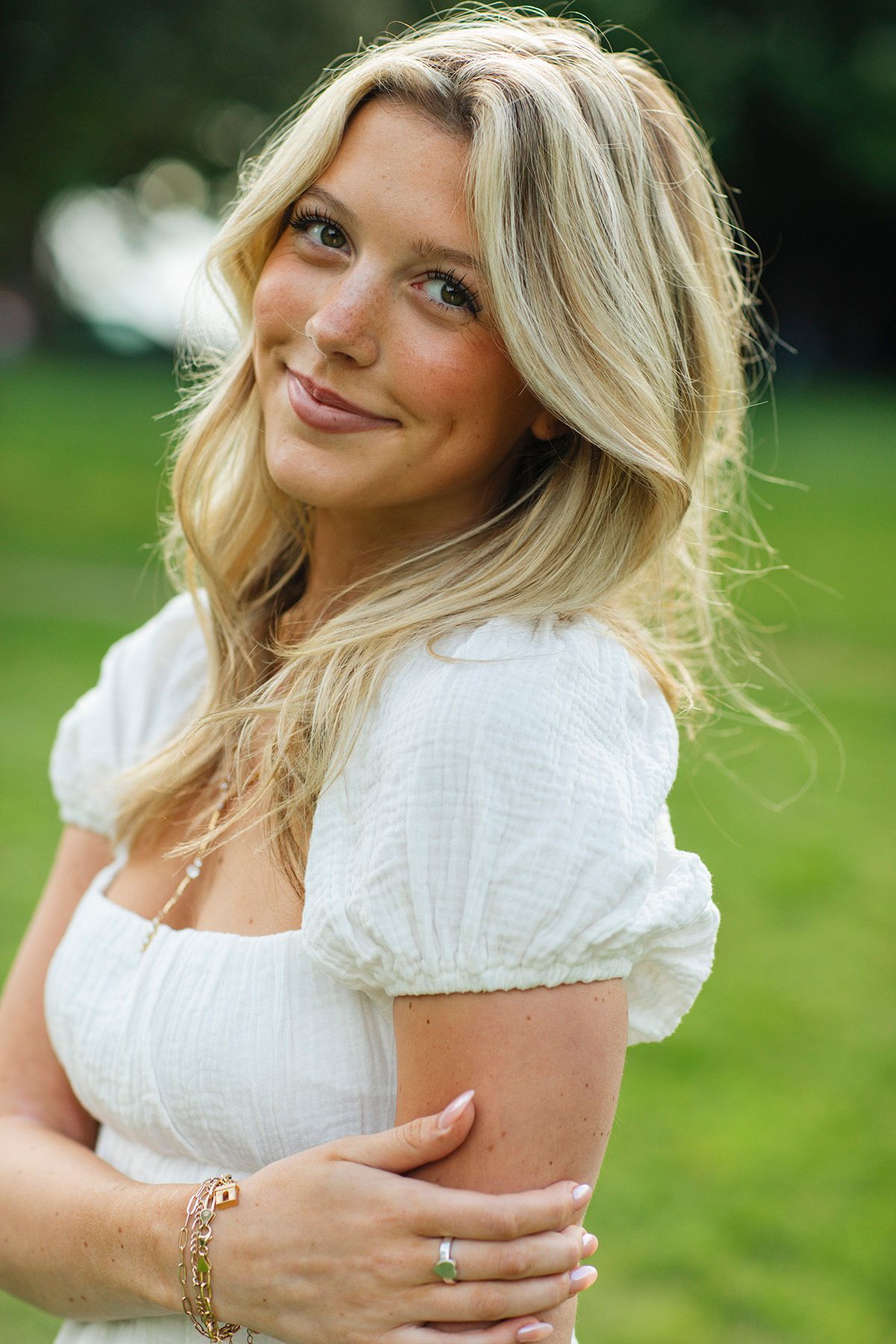 Smiling blonde woman in white puff-sleeve dress posing outdoors in a green park setting.