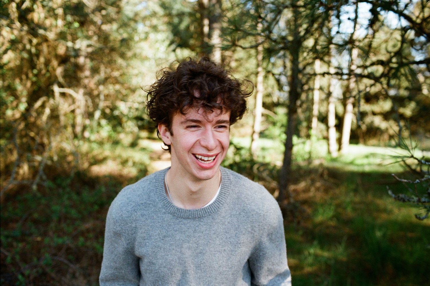 Young man with curly hair smiling outdoors in a forest setting, wearing a gray sweater.