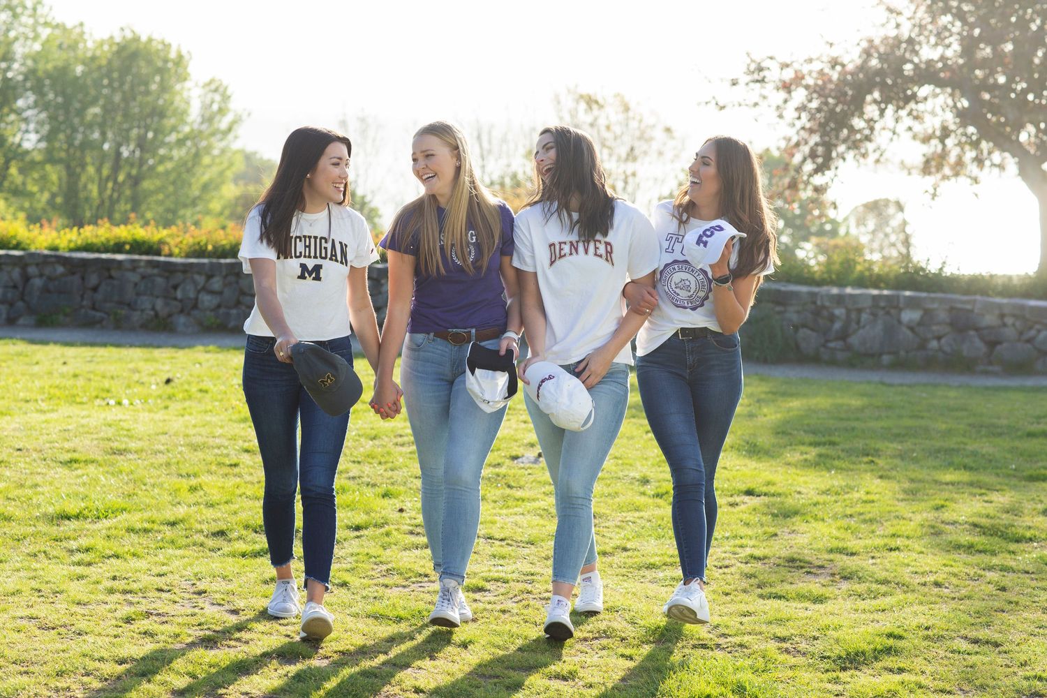 Four young women in college shirts walk together on a sunny green lawn, smiling and holding helmets.