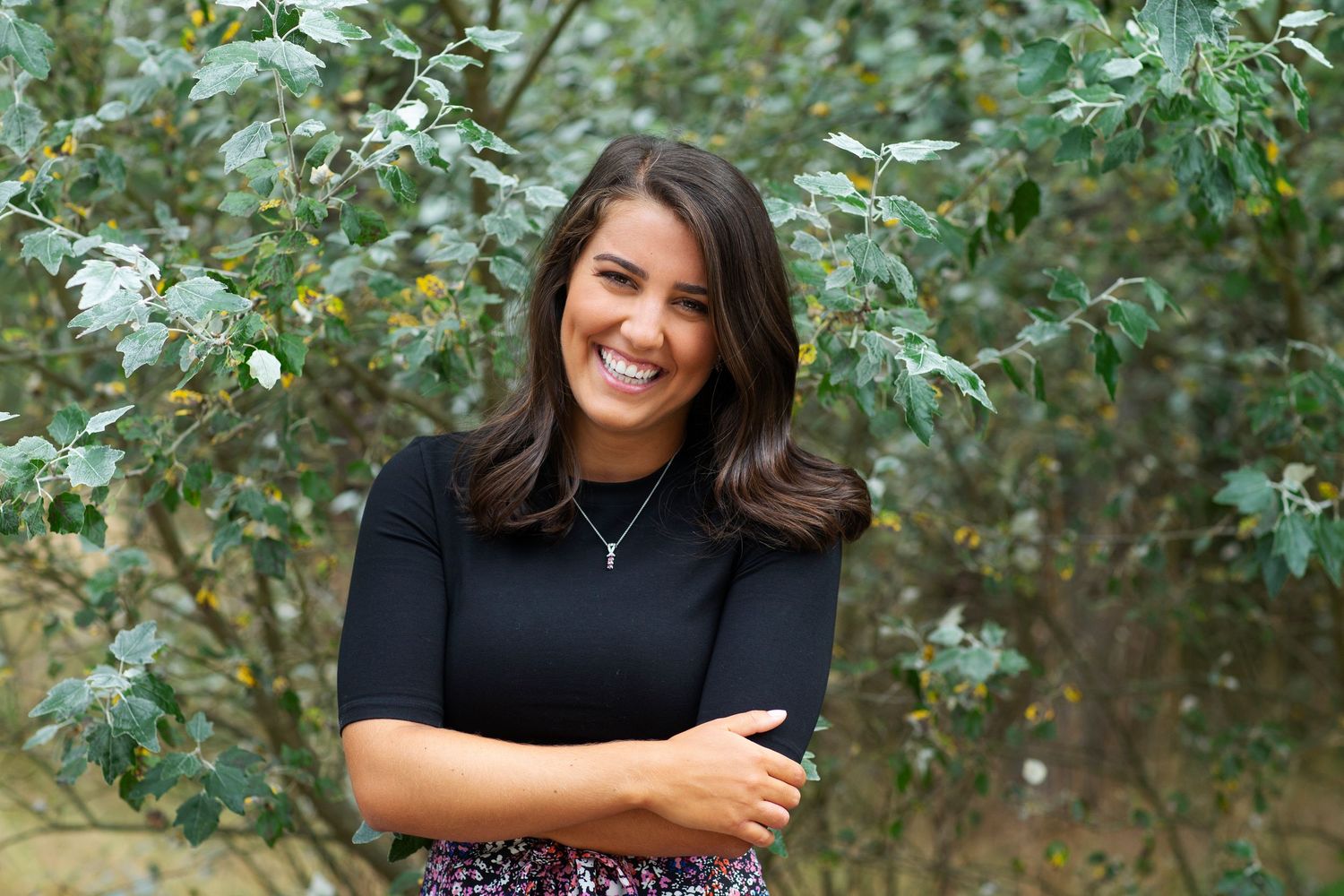 Cheerful woman in black top with arms crossed near green bushes.