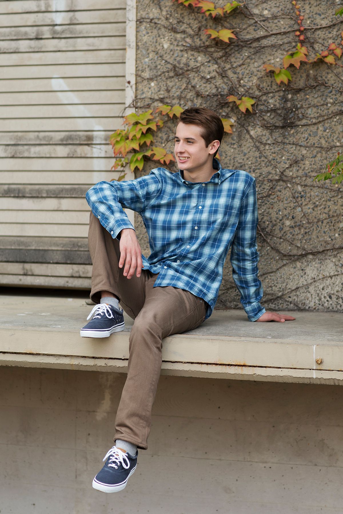 Young man in blue plaid shirt and khakis sits on concrete ledge, smiling outdoors near ivy-covered stone wall.