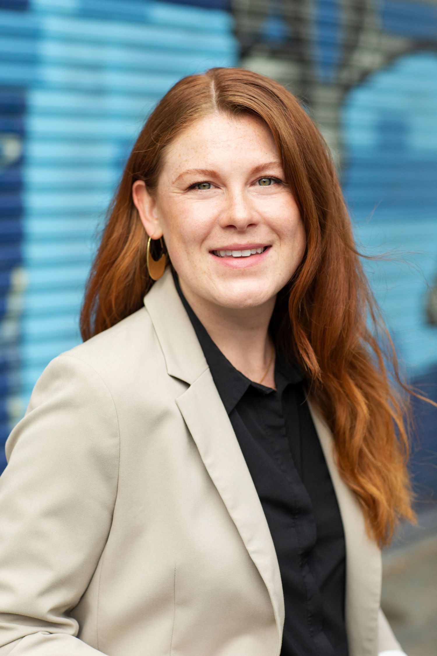 Redhead woman in beige blazer smiling outdoors against blue wall.