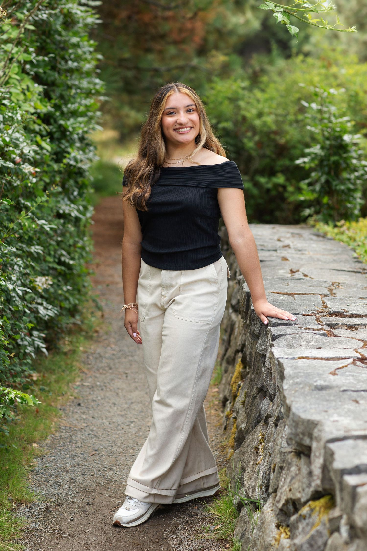 Smiling young woman in black off-shoulder top and white wide-leg pants posing on a stone garden path.