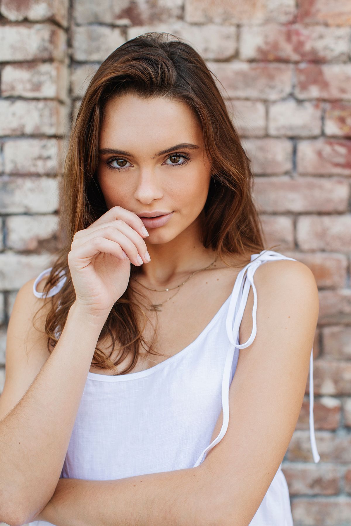 Young woman with brown hair in white sundress poses thoughtfully against rustic brick wall.