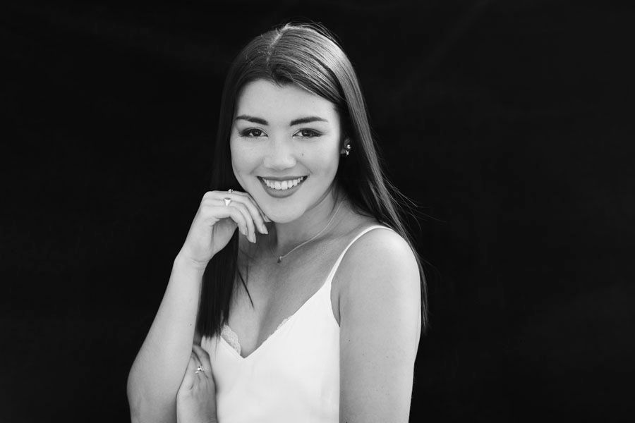 Black and white portrait of a smiling young woman with long hair wearing a white dress against dark background.