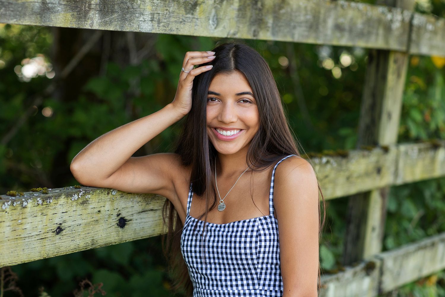 Young woman in gingham dress smiling outdoors by wooden fence with lush green foliage background.