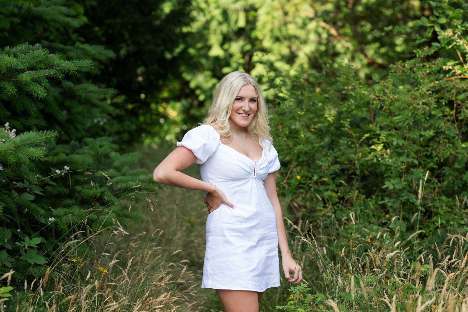 Blonde woman in a white mini dress posing among lush green trees and tall golden grass outdoors.