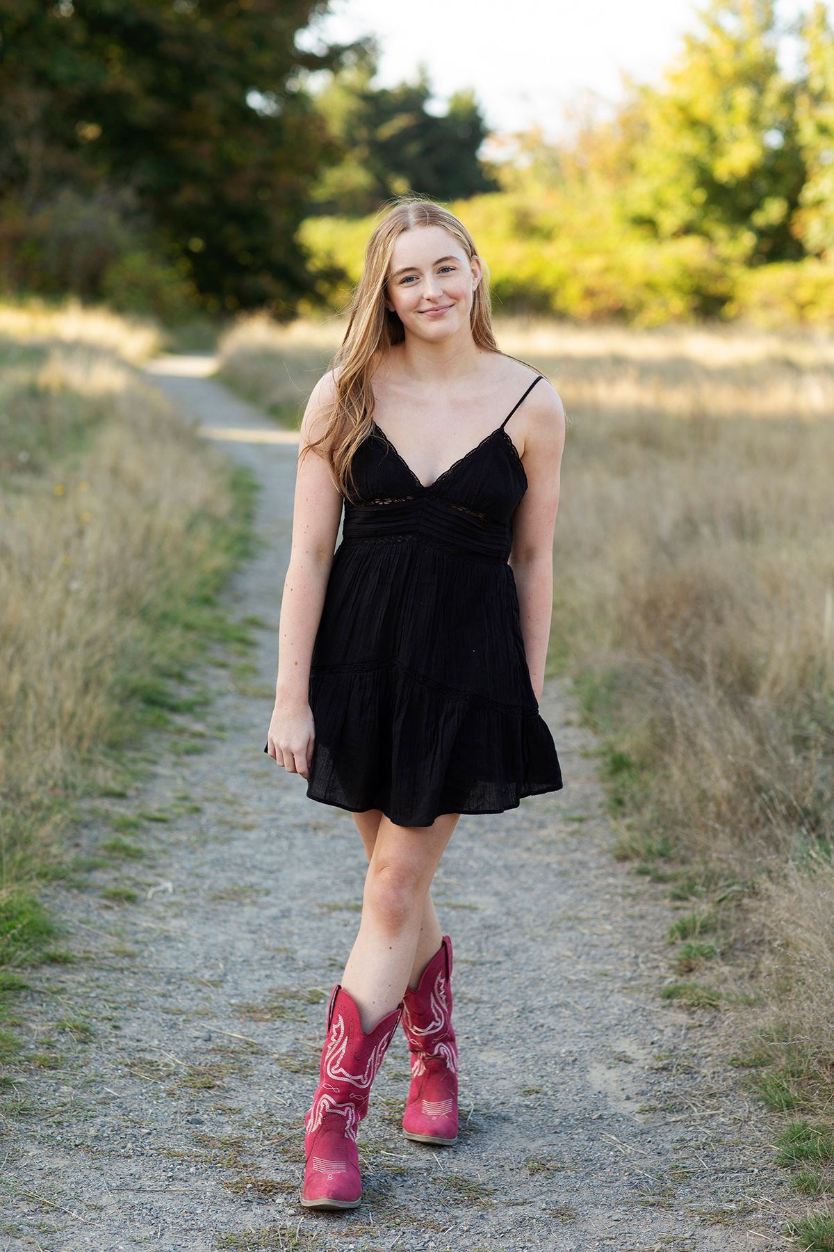 Young woman in black sundress and pink cowboy boots smiling on a rustic dirt path surrounded by golden fields.