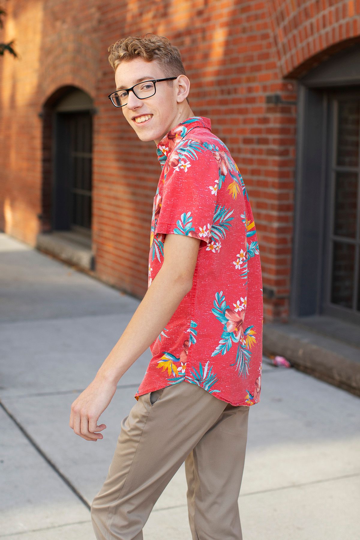 Young man in red floral Hawaiian shirt and khaki pants smiling while walking on a city sidewalk.