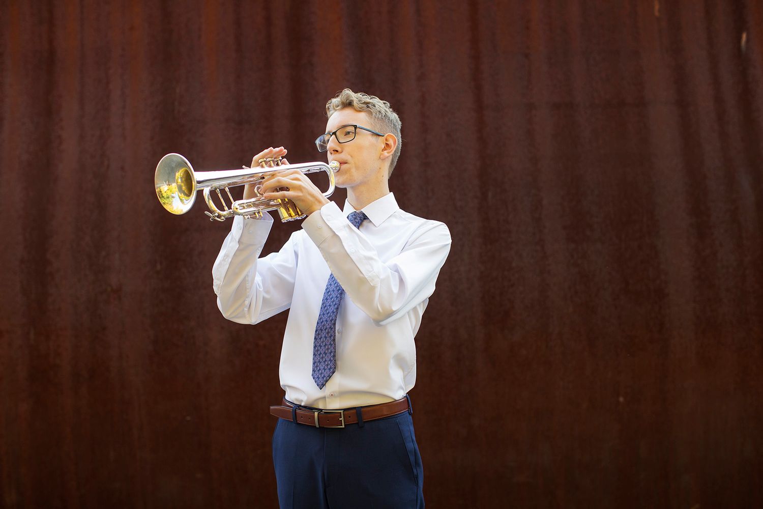 Man in white shirt and tie playing trumpet outdoors against a rustic metal wall backdrop.