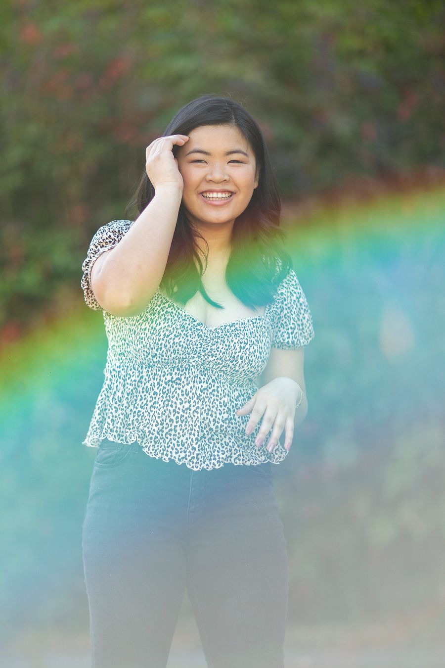 A smiling young Asian woman in a floral top poses outdoors with a vibrant rainbow arc behind her.