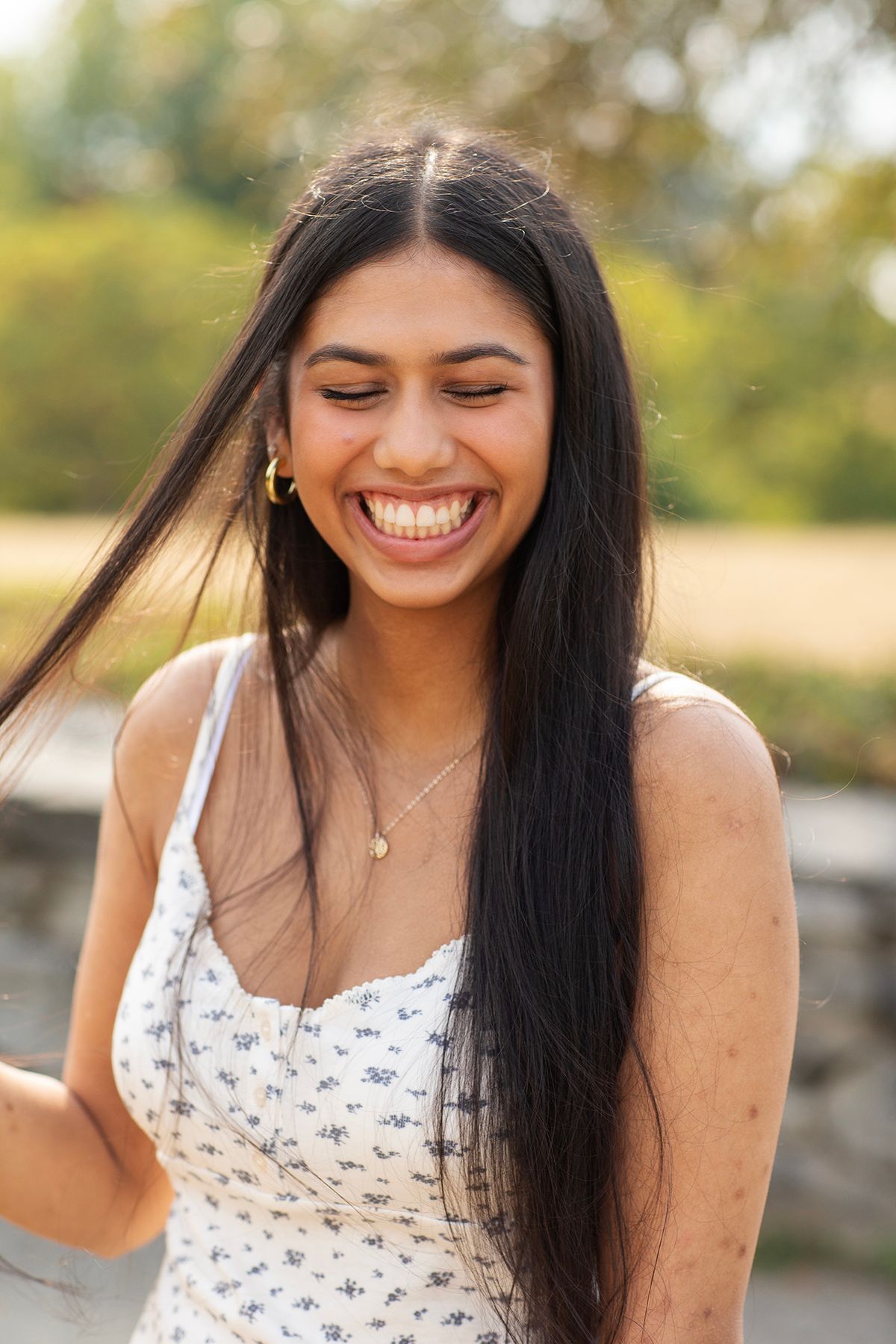 Joyful young woman with long dark hair laughing outdoors in white floral dress with bokeh background.