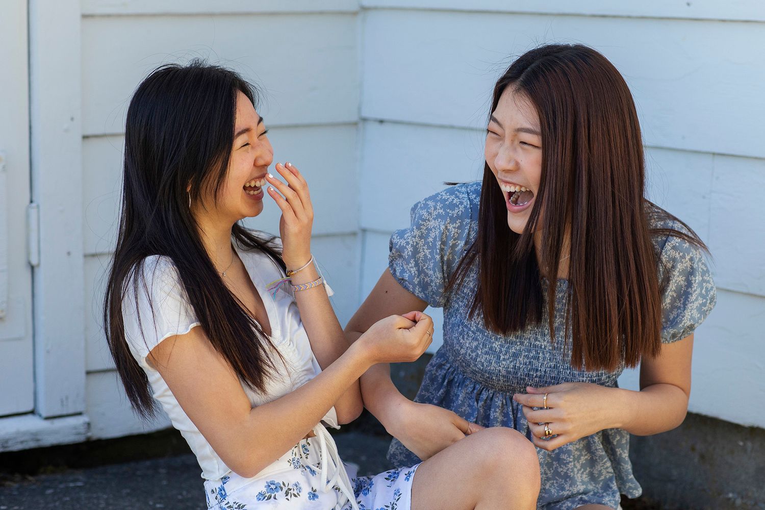 Two young women laughing and sharing a joyful moment together outdoors, sitting against a white wooden wall.