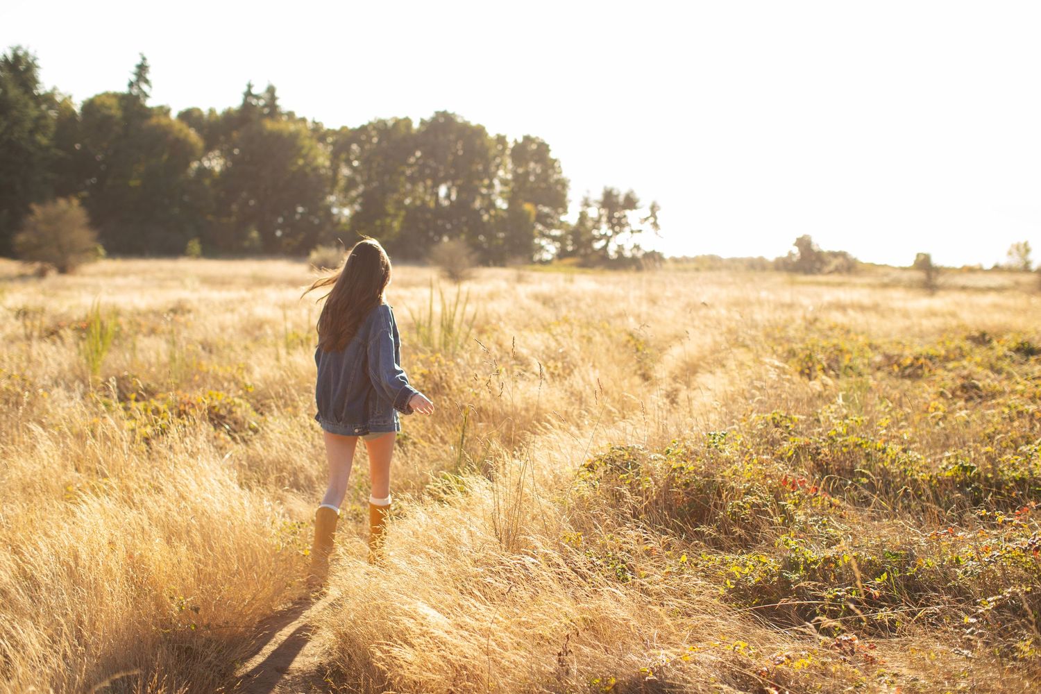 Young woman walking through golden sunlit field wearing blue shirt and yellow boots.