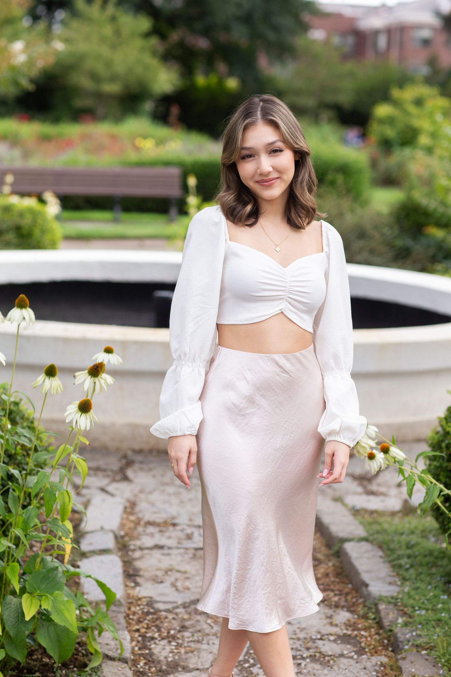 Smiling young woman in a white crop top and blush satin midi skirt posing in a garden park.