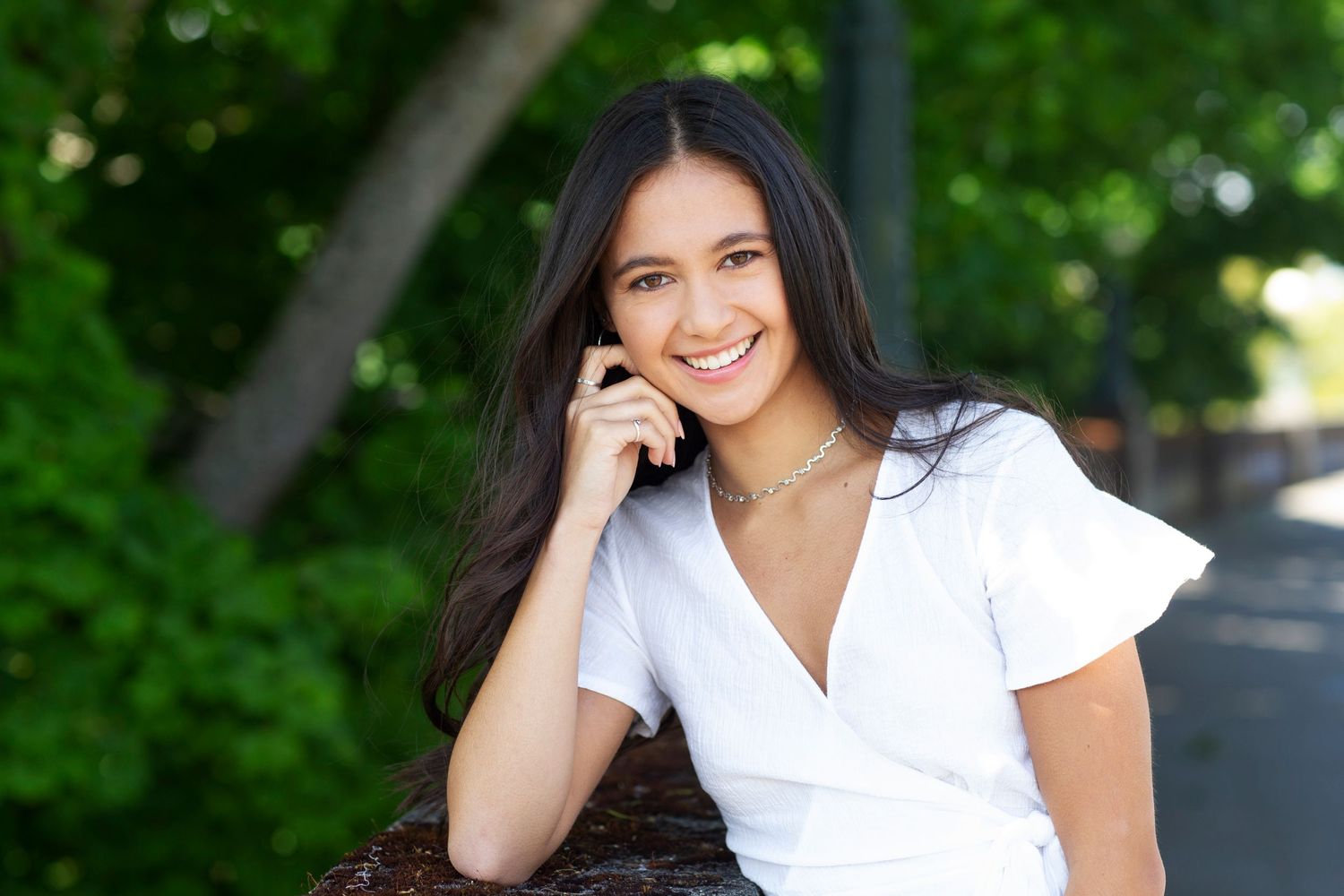 Smiling young woman with dark hair in white dress posing outdoors surrounded by green trees.