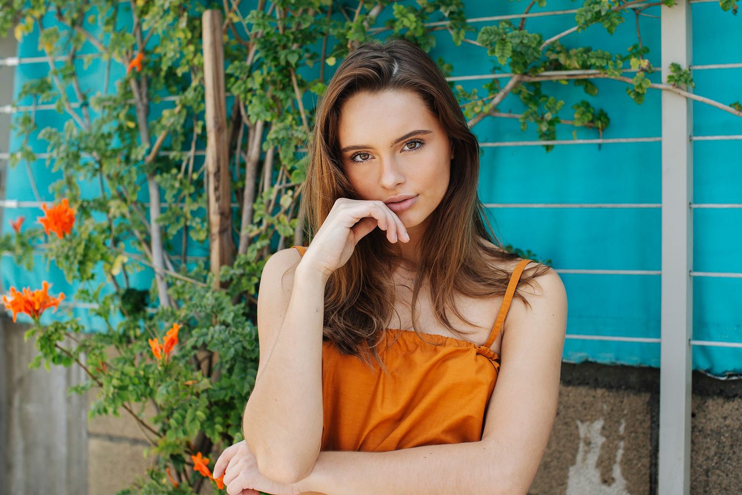 Young woman in orange dress posing thoughtfully against turquoise wall with green vines and orange flowers.