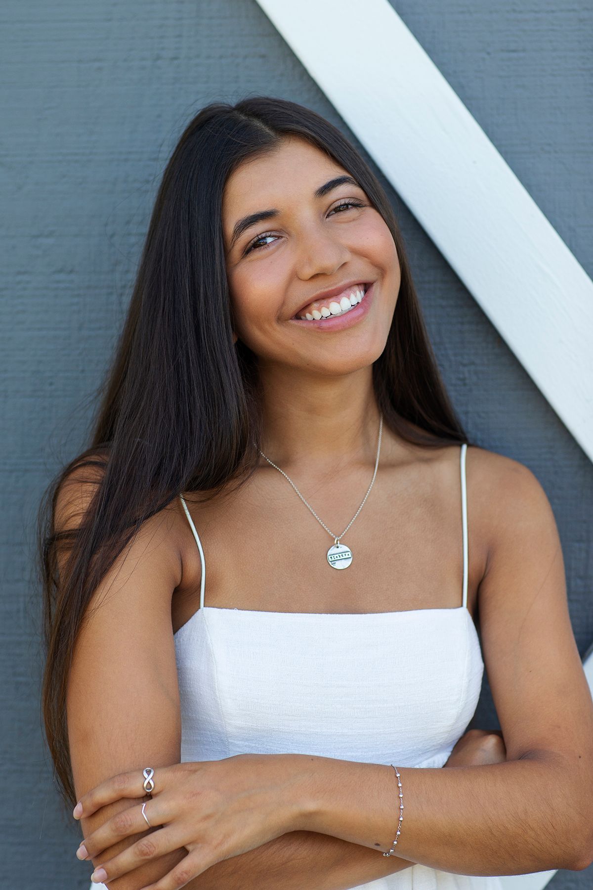 Smiling young woman with long dark hair wearing a white dress and silver necklace, posing against a gray wall.