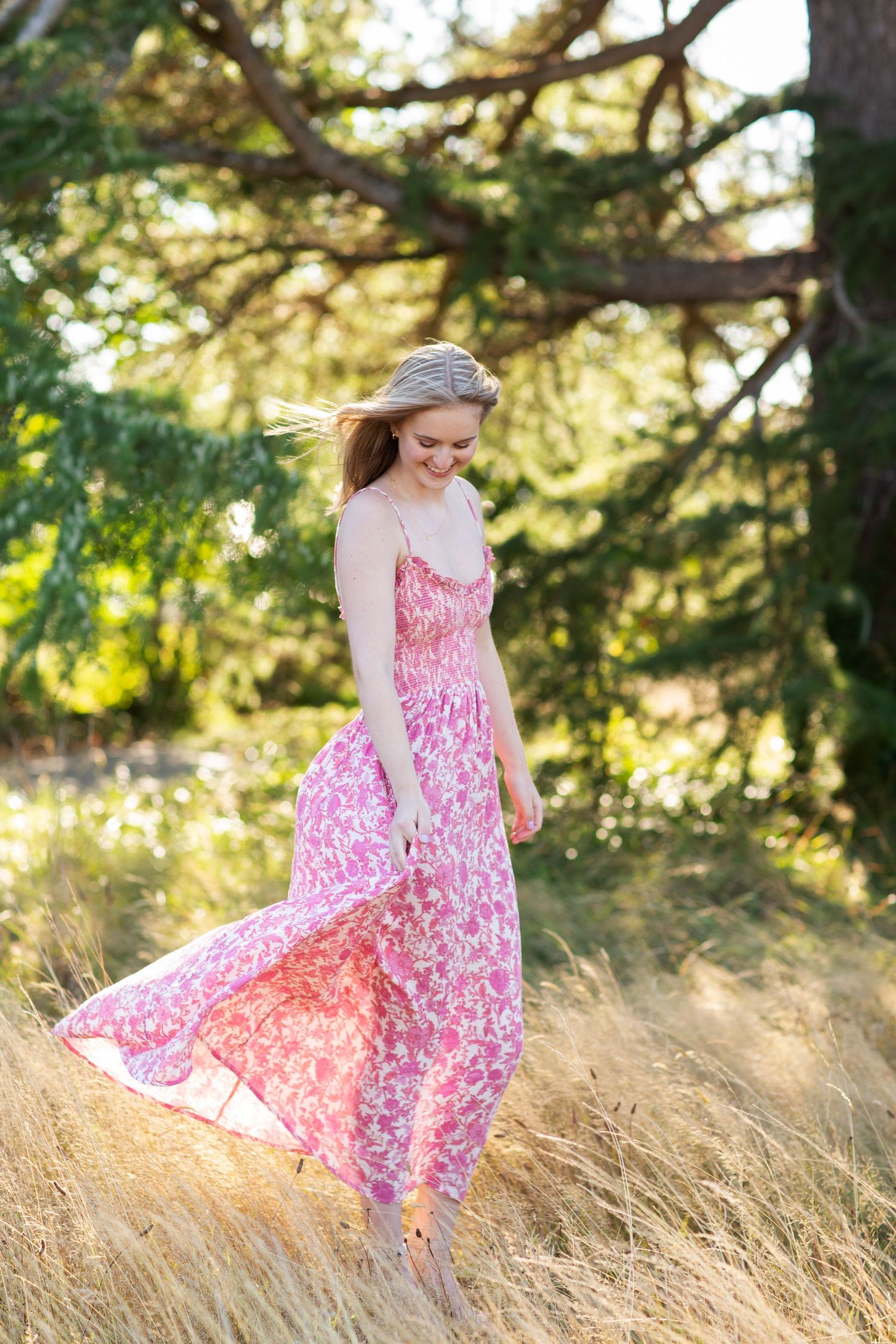 Blonde woman in flowing pink floral maxi dress walking through golden grass near pine trees.