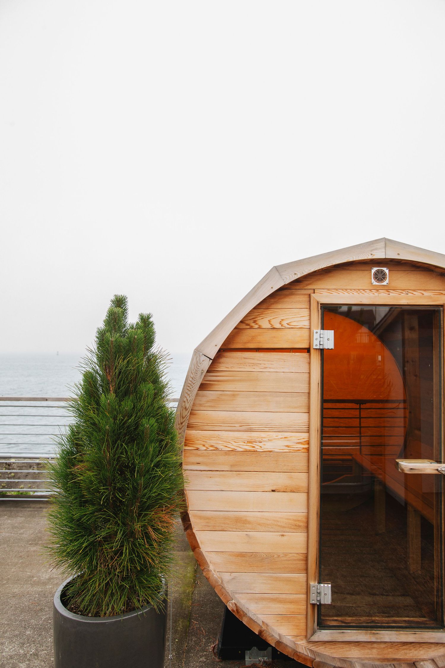 Barrel sauna with wooden exterior and open door beside a potted evergreen tree on an outdoor deck overlooking the sea.