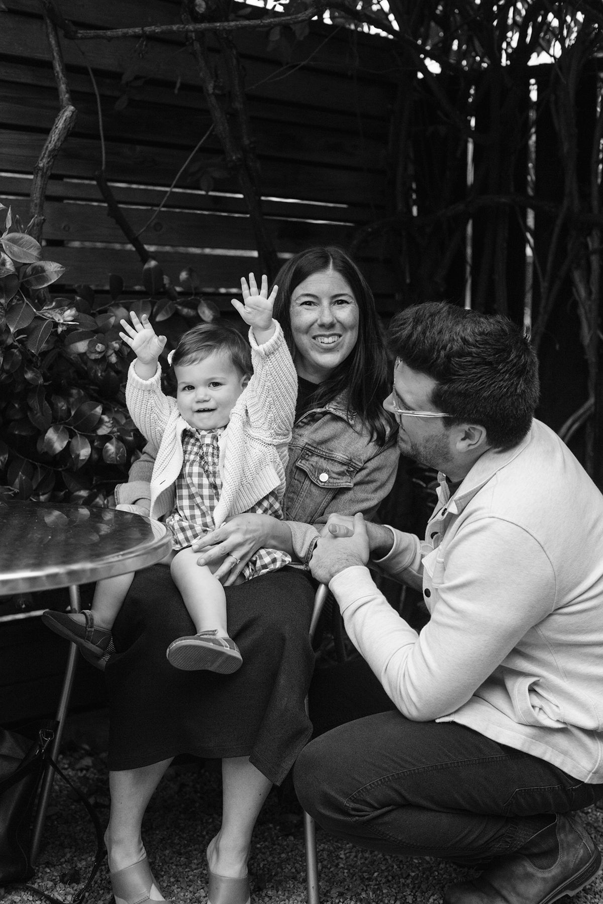 Black and white photo of a happy family with a baby waving hands, sitting outdoors at a table.