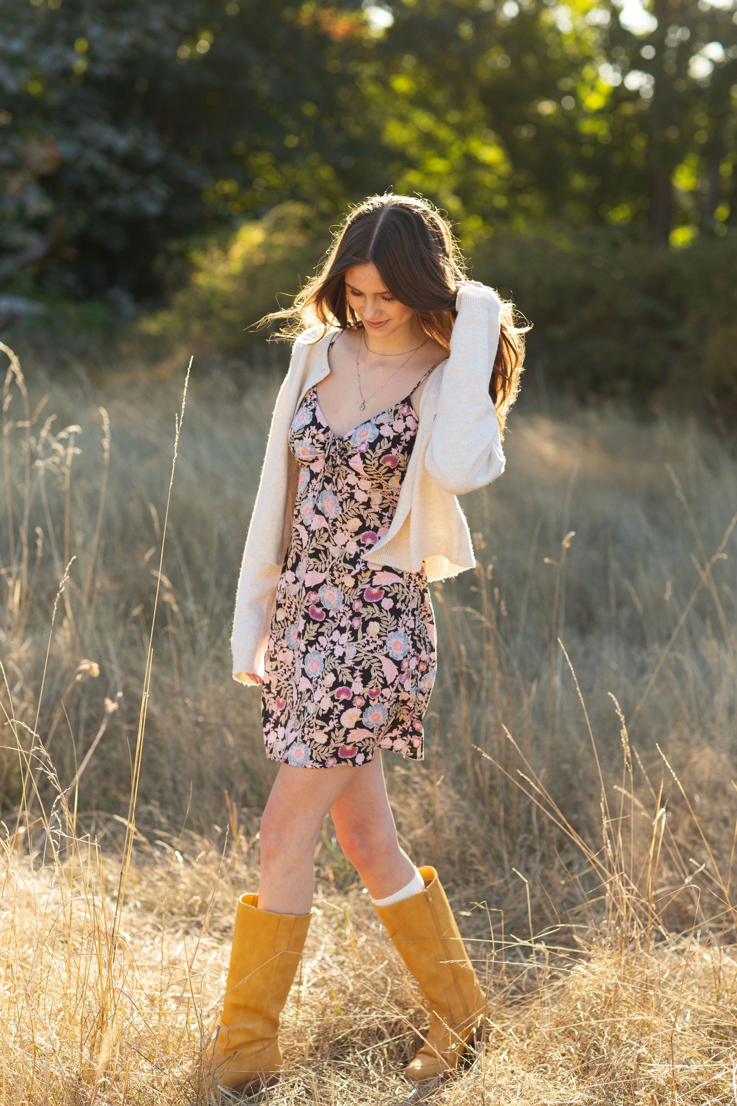 Woman in floral mini dress, white cardigan, and tan boots walks through sunlit dry grass field.