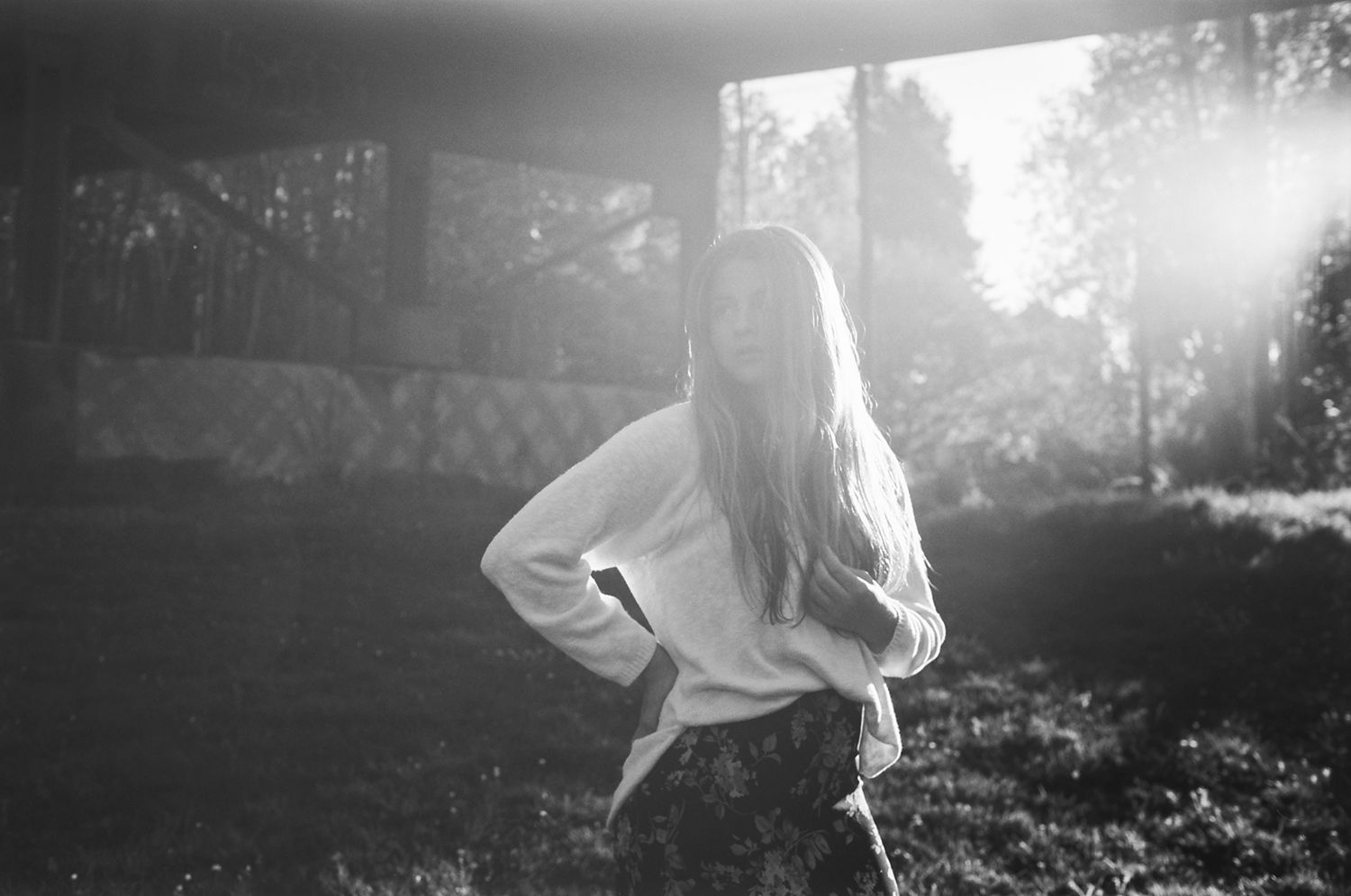 Black and white photo of a young woman with long hair posing outdoors in sunlight wearing a white sweater.