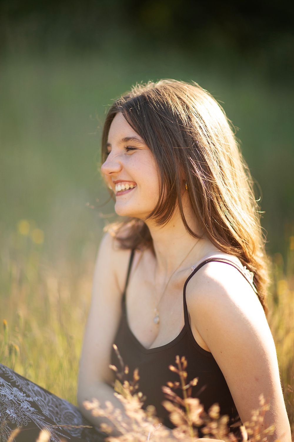 Young woman with long brown hair smiling outdoors in golden sunlight, wearing a black top in a grassy field.