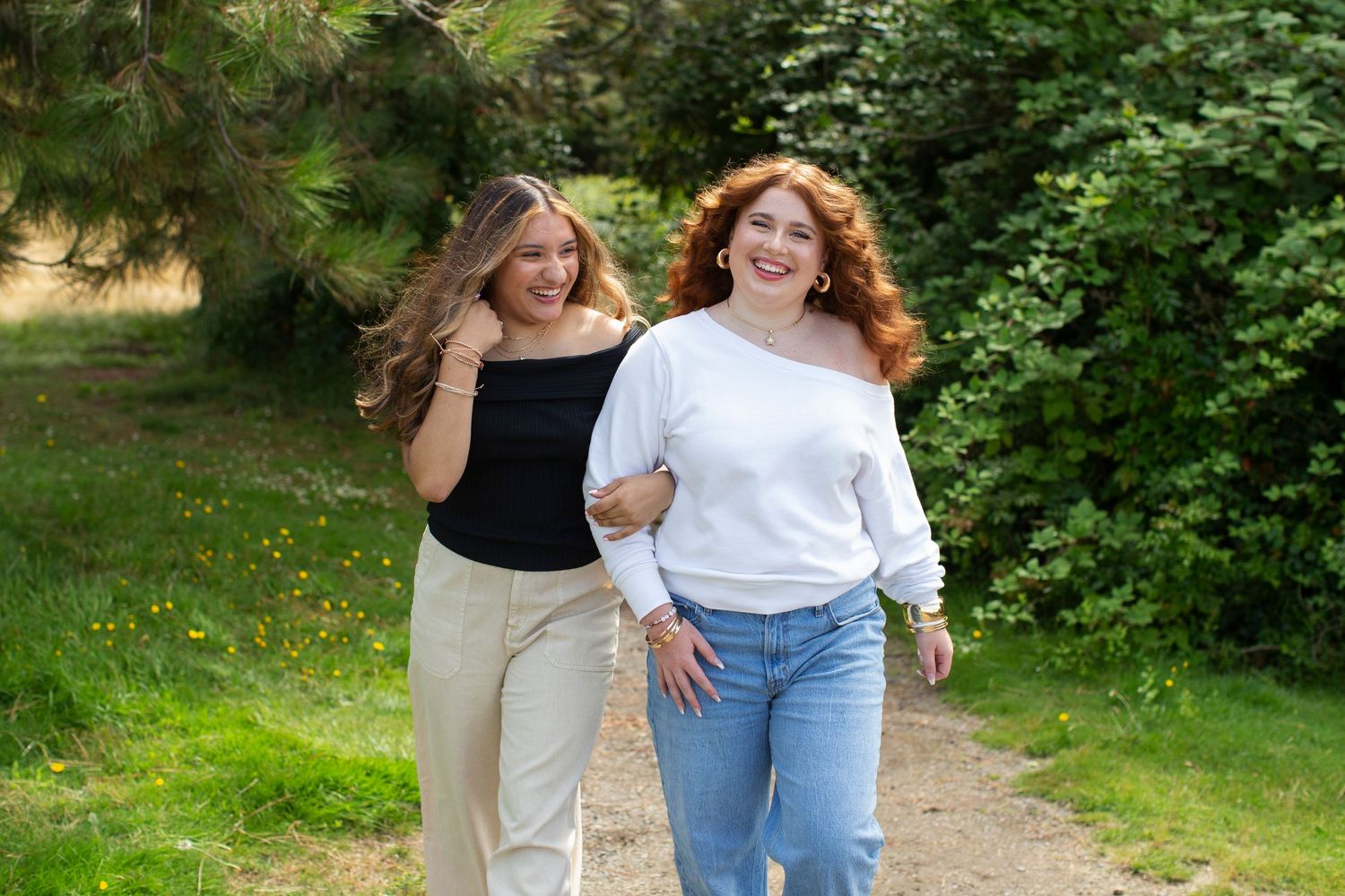 Two young women smiling and walking together on a garden path surrounded by lush green trees.