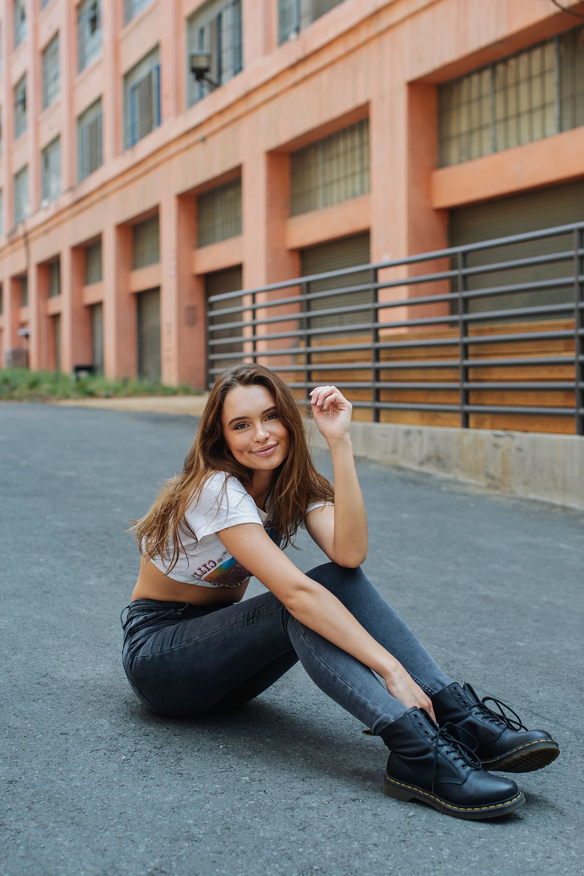 Smiling young woman in white crop top and jeans sitting on pavement near urban building.