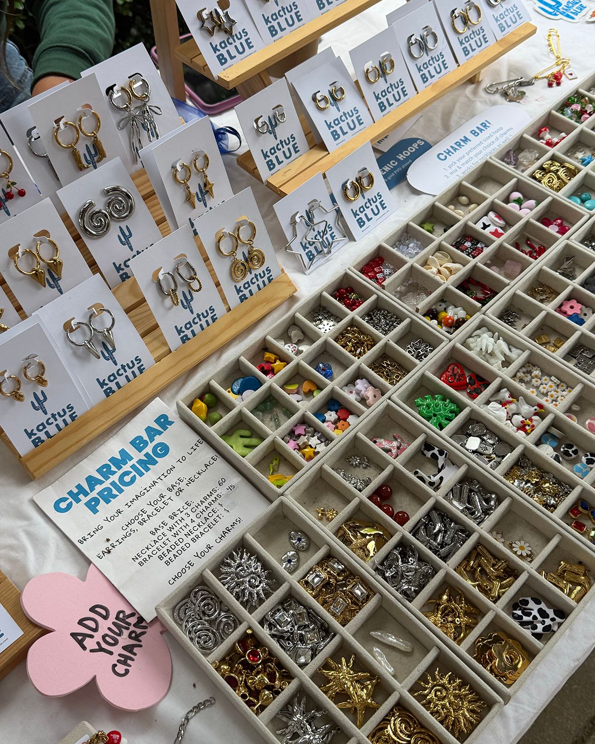 A craft market display of Kactus Blue earrings on wooden stands beside a charm bar with sorted beads and charms in a divided tray.