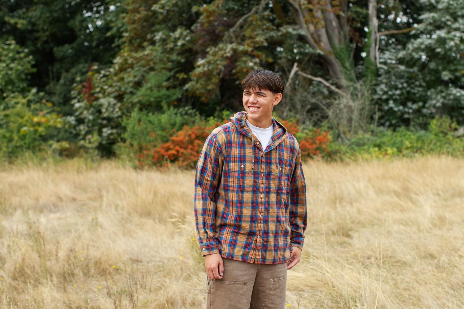 Young man in plaid flannel shirt smiling in golden field with autumn trees.