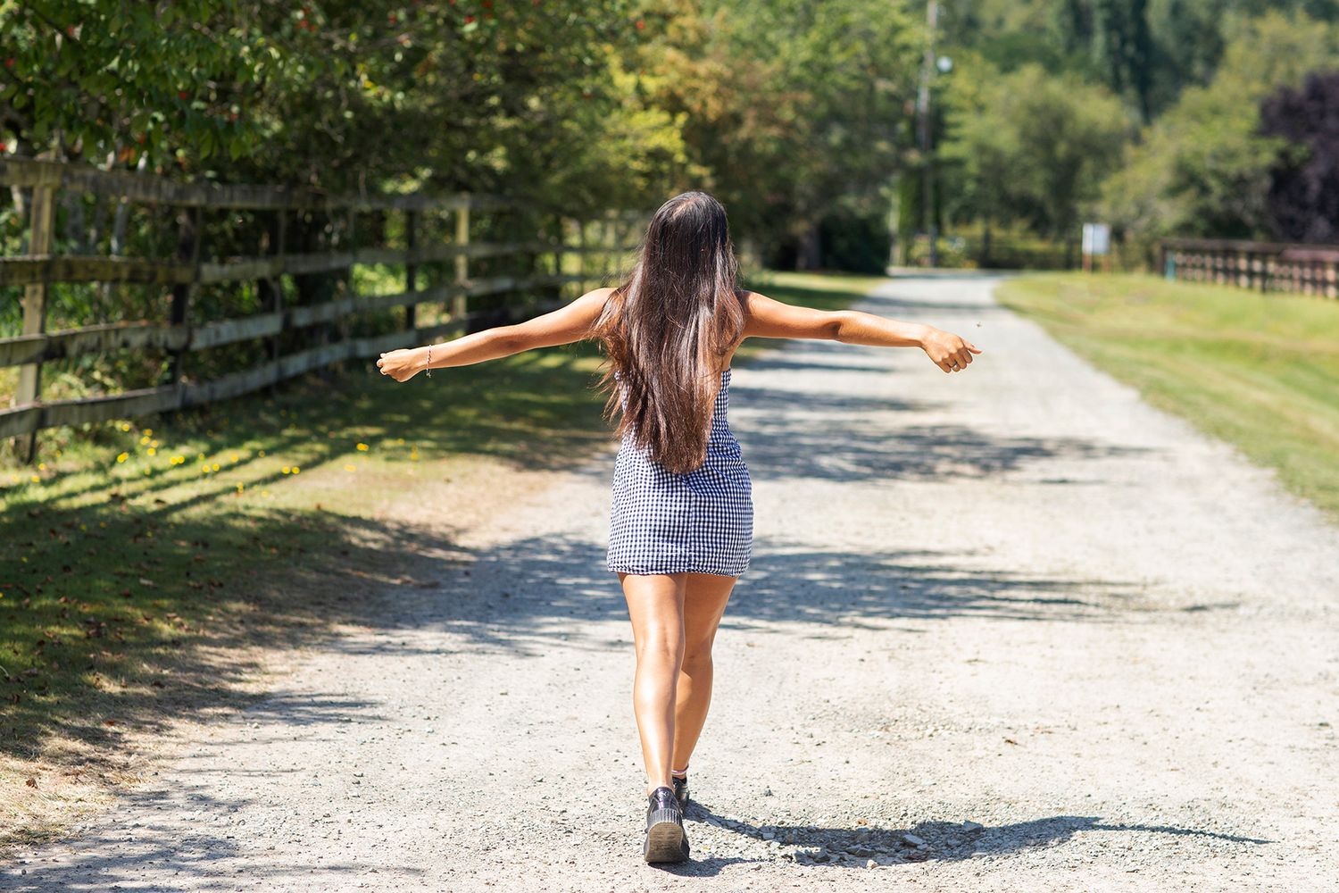 Young woman in shorts walking down a sunny gravel path with arms outstretched, surrounded by green trees and a wooden fence.