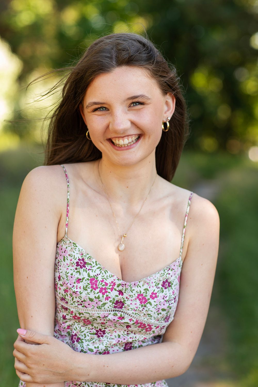 Smiling young woman with long brown hair wearing a floral sundress outdoors in a green park setting.