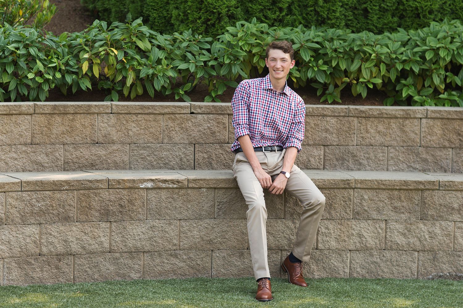 Young man in plaid shirt and khakis sitting on a stone retaining wall outdoors, smiling in a garden setting.