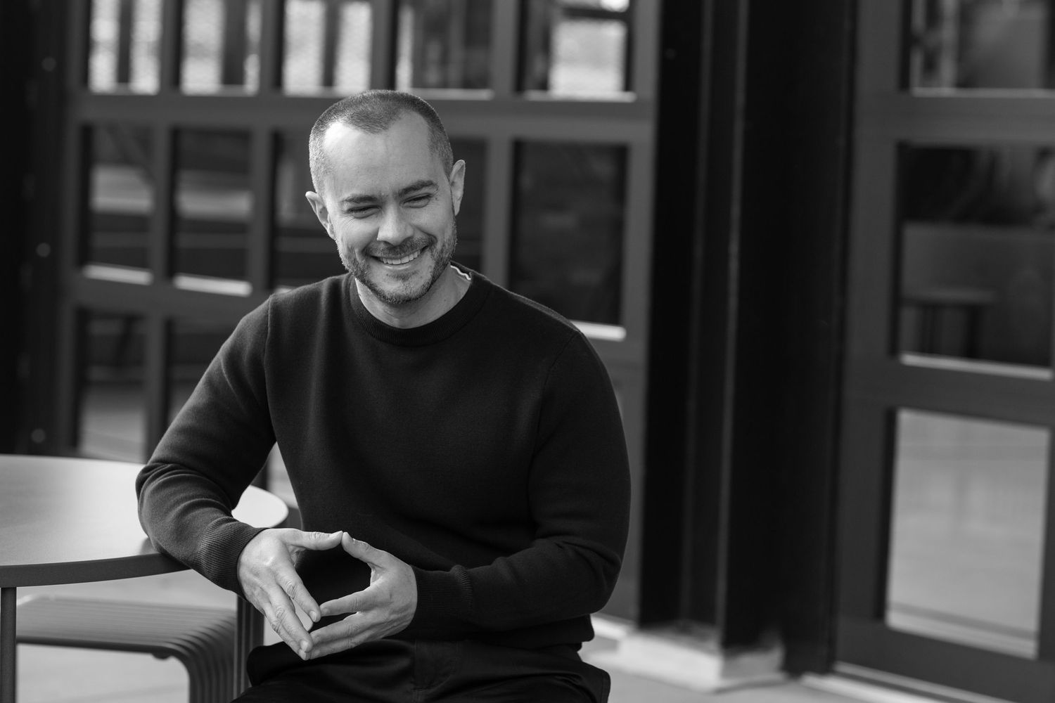 Smiling bald man in dark sweater sitting relaxed in black and white portrait.
