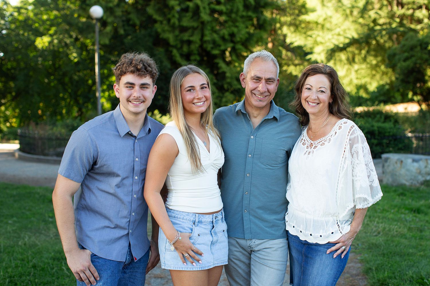 A smiling family of four poses outdoors in a lush green park, dressed in casual summer attire.