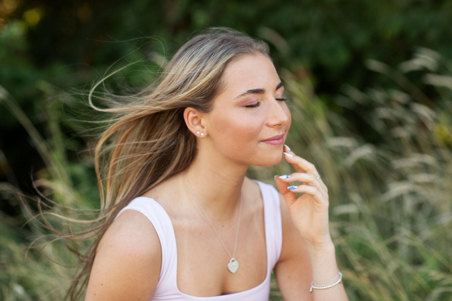 Young woman with long blonde hair enjoys a serene outdoor moment, eyes closed, touching her lips gently.