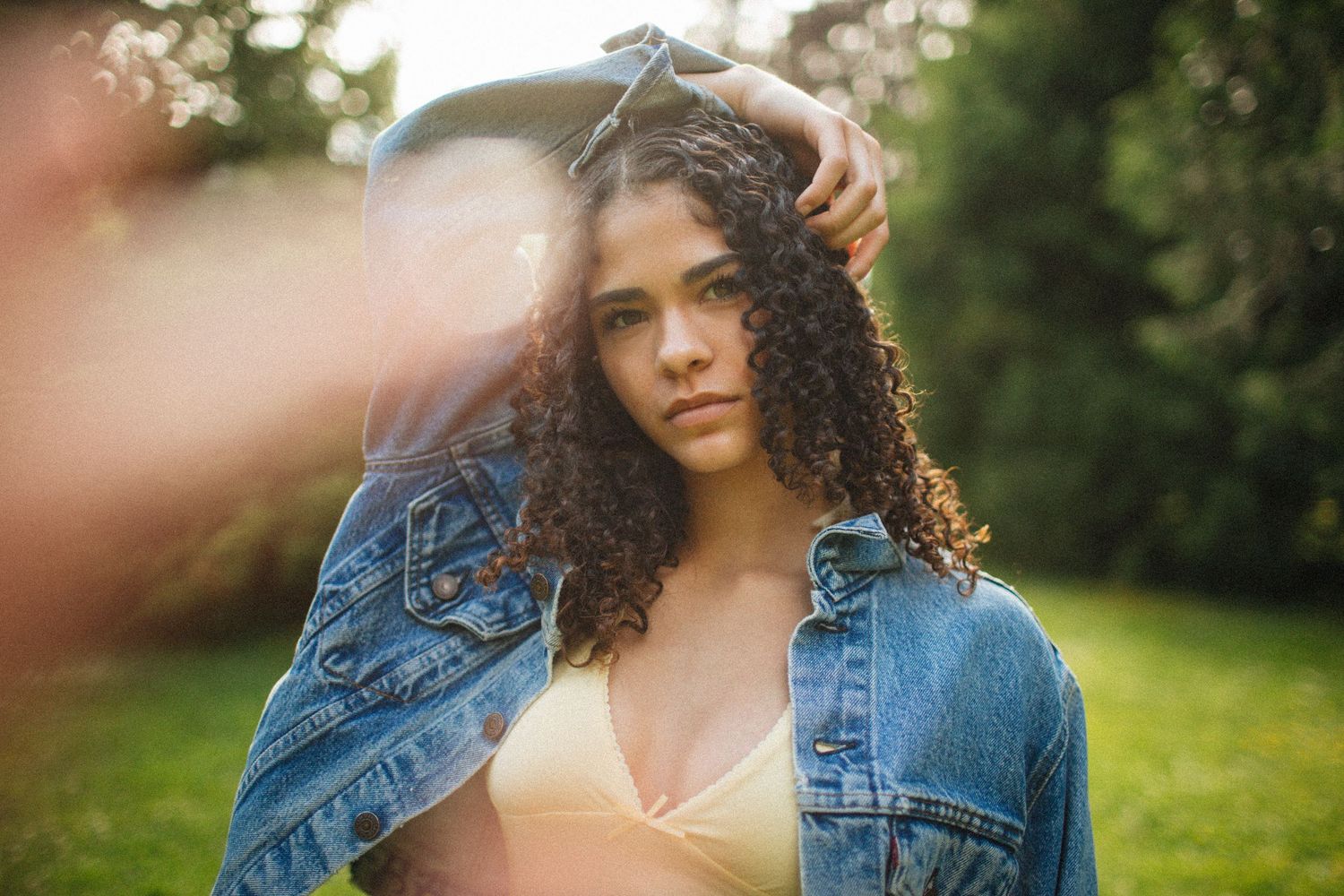 Young woman with curly hair in denim jacket poses outdoors in golden sunlight with bokeh effect.