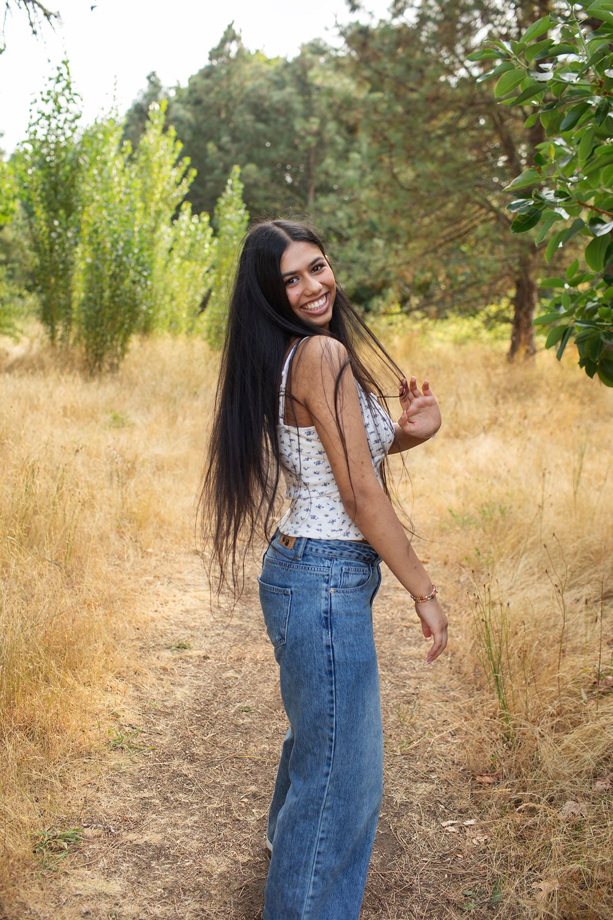 Happy young woman in jeans and floral top walking on forest trail looking back over shoulder.