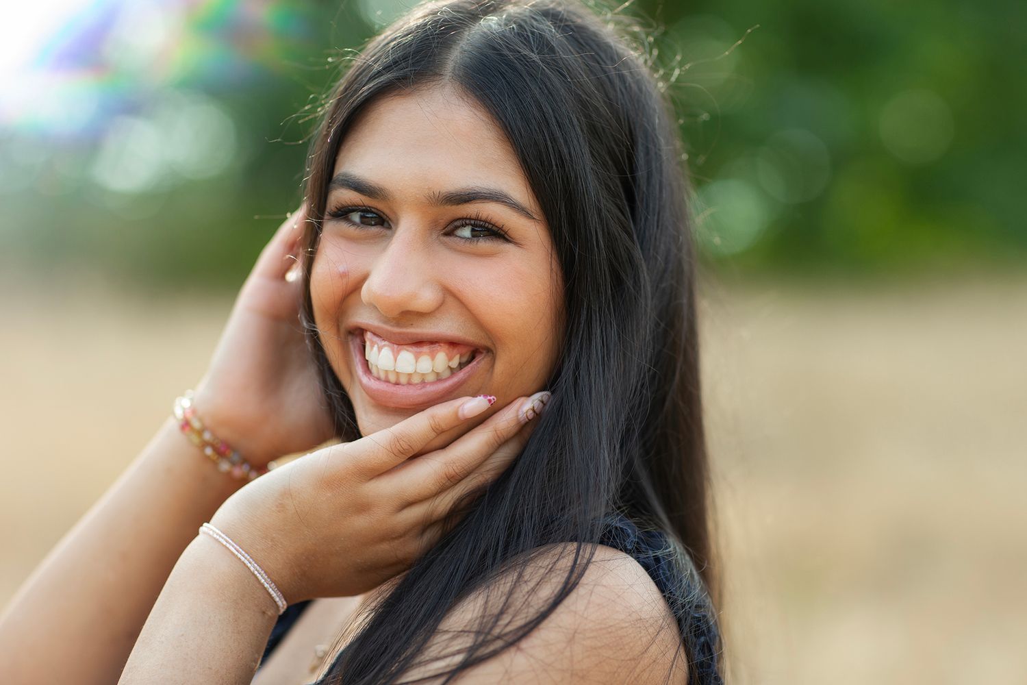Close-up portrait of smiling young woman with long dark hair and bracelets outdoors.