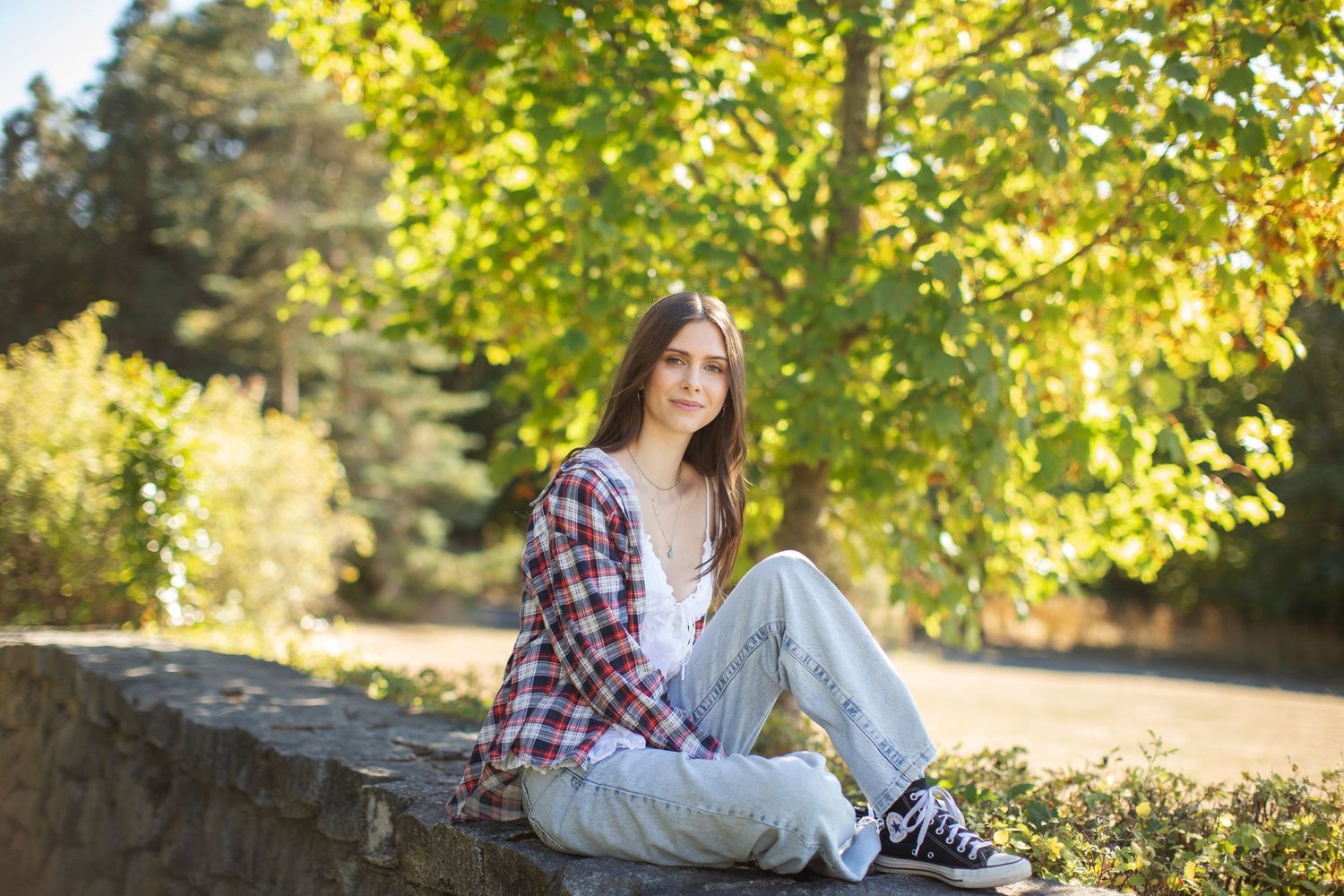 Young woman sitting on stone wall in park wearing plaid shirt, jeans, and black sneakers.