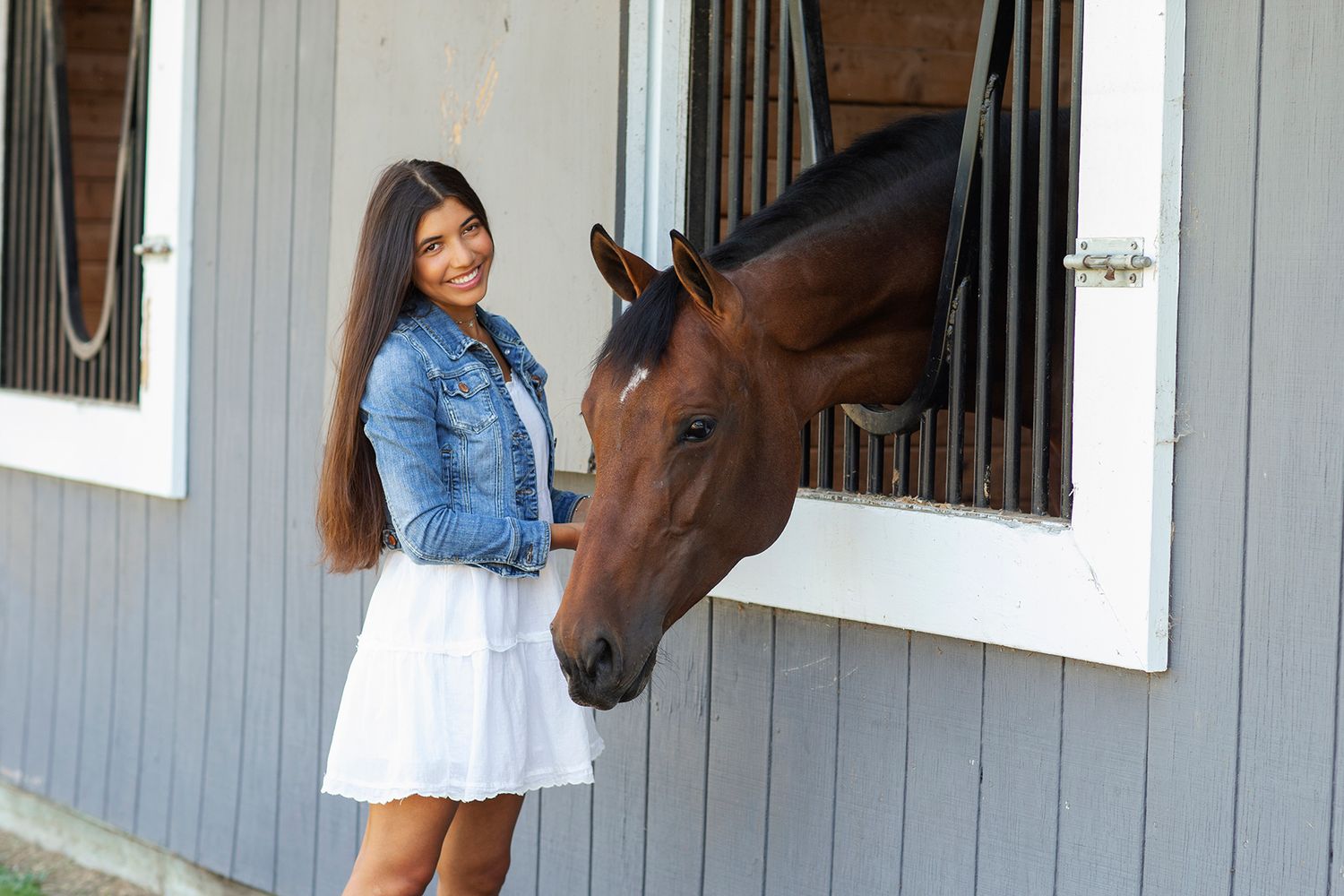 Young woman in denim jacket and white skirt smiling beside a brown horse at a stable window.