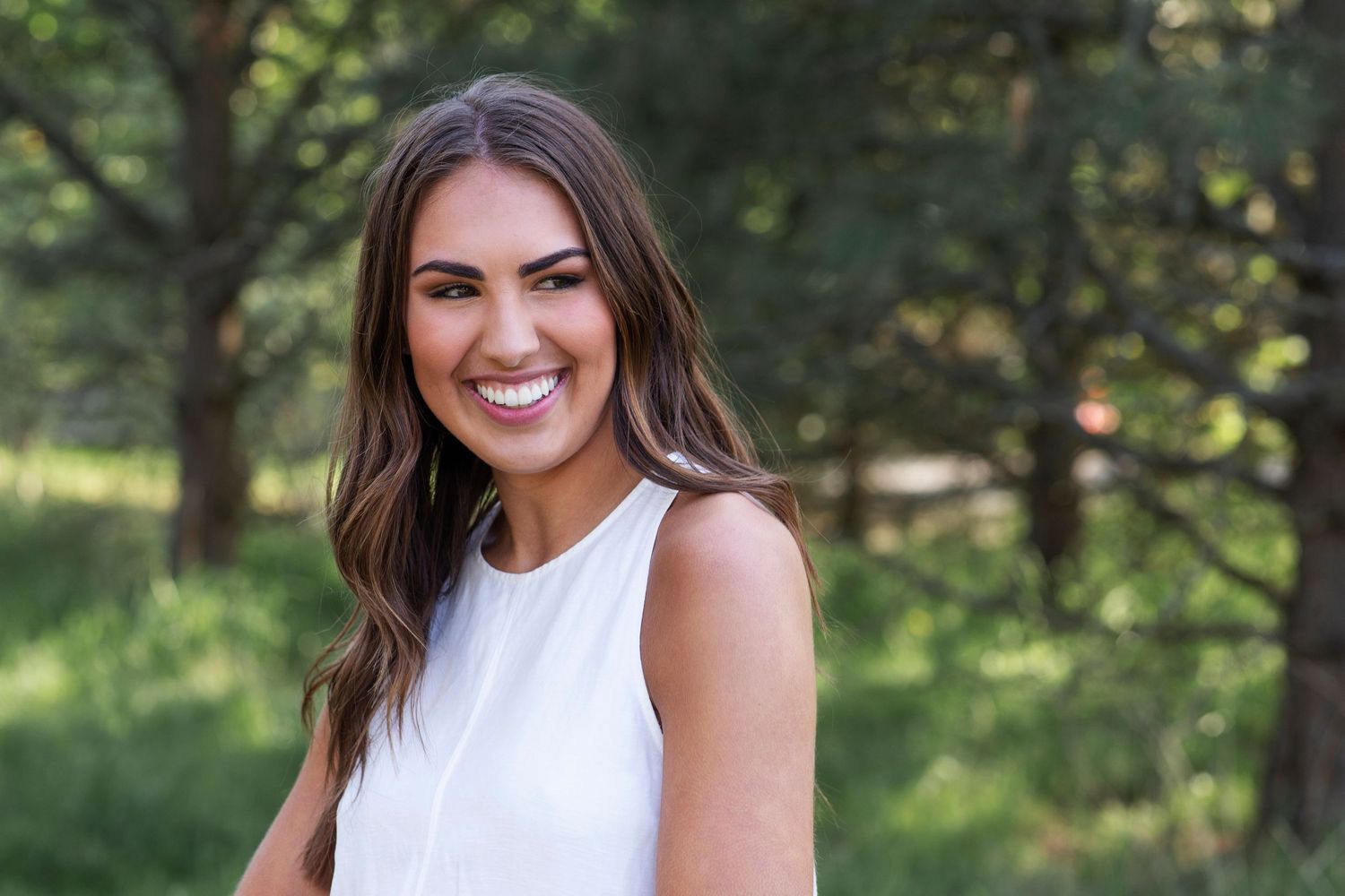Smiling young woman with long brown hair wearing a white sleeveless top, posing outdoors with trees in background.