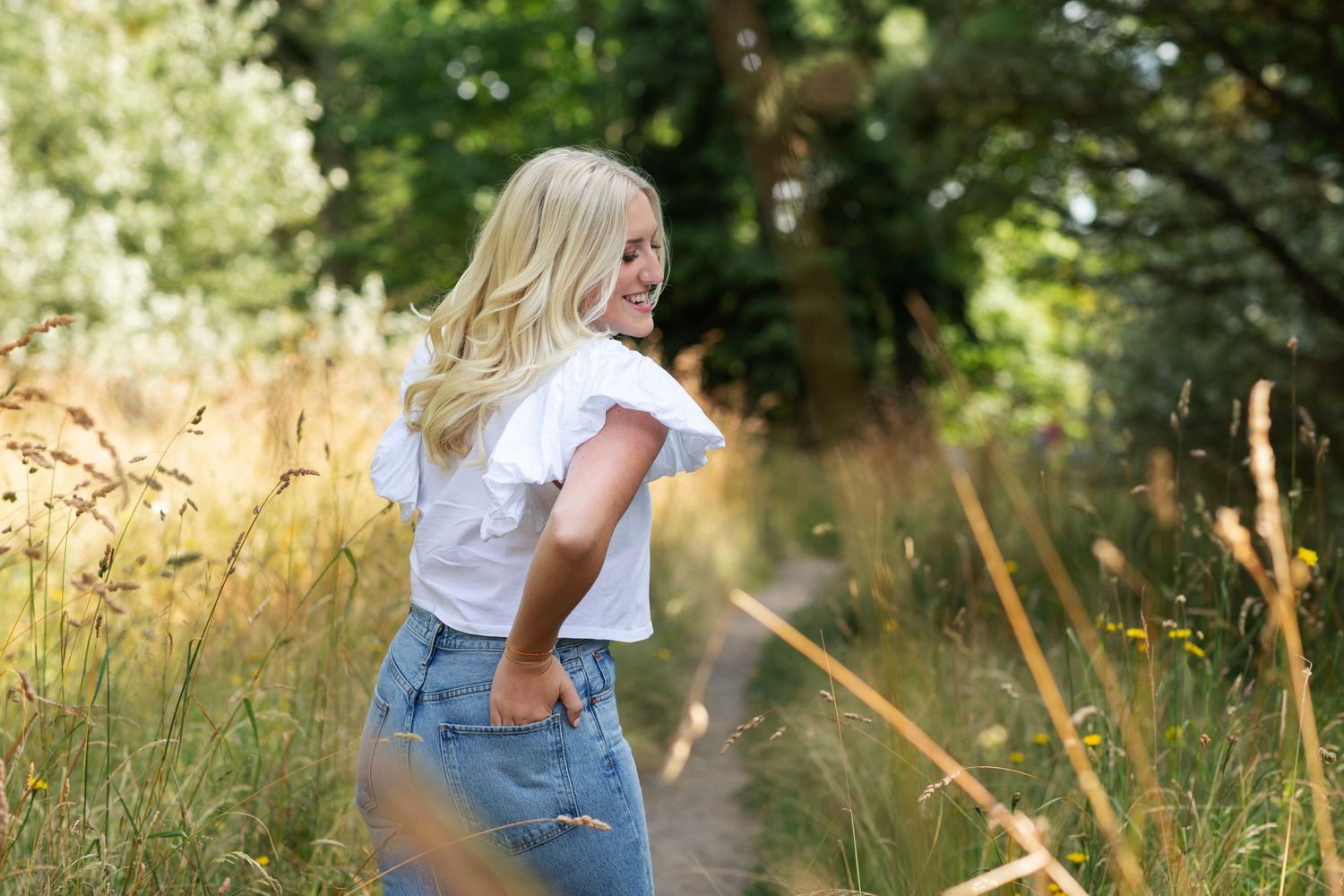 Blonde woman in white crop top and jeans smiling while walking along a sunny nature trail.