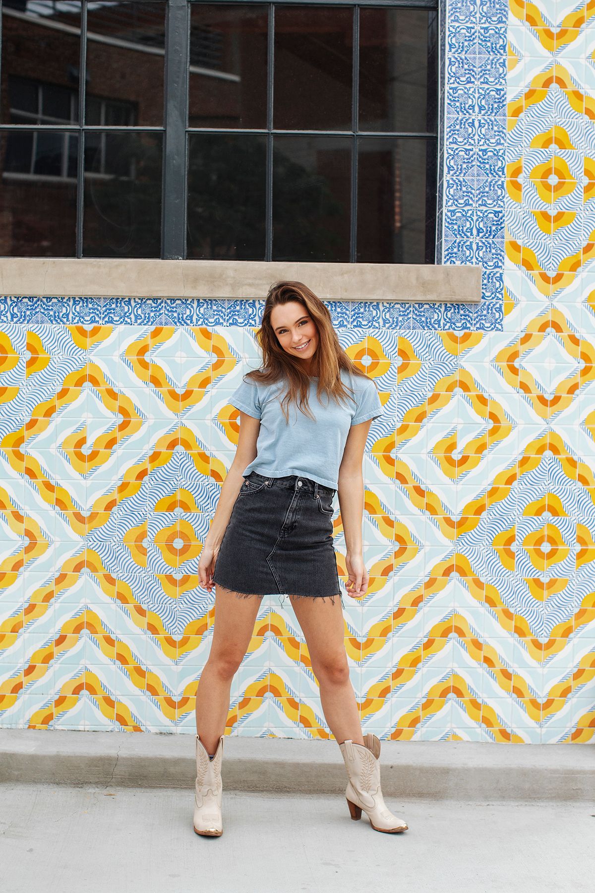 Young woman in blue tee, denim mini skirt, and cowboy boots posing against a colorful geometric tiled wall.
