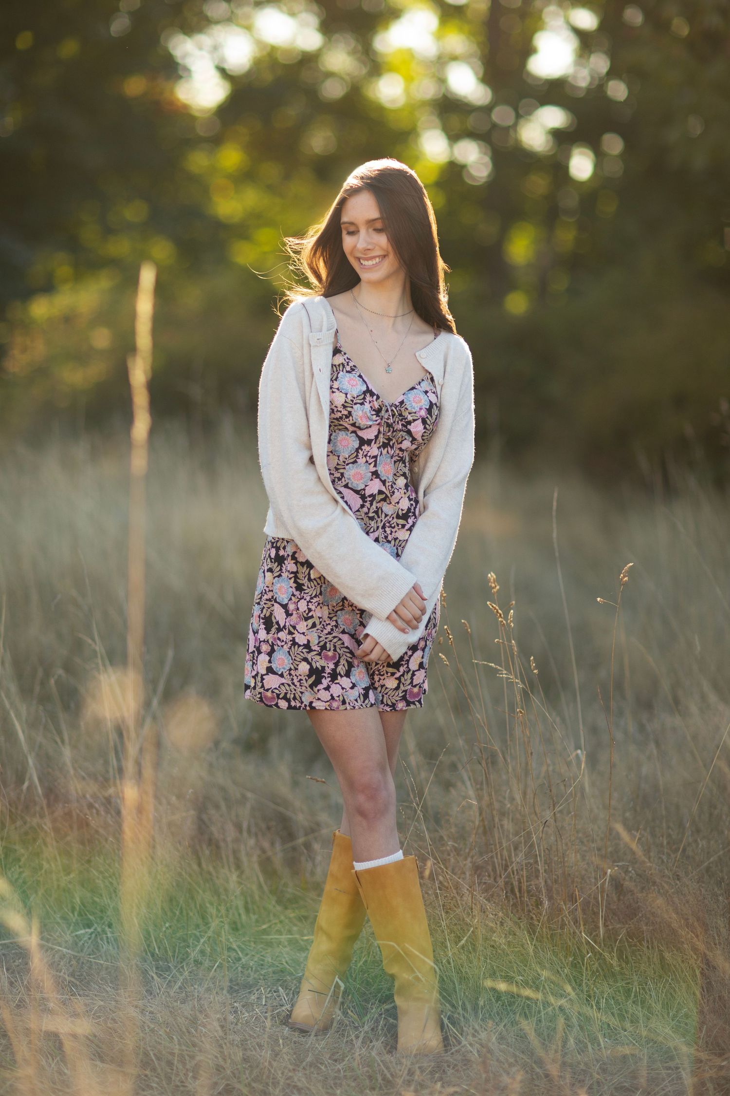 Woman in floral mini dress and tan boots smiles while standing in golden sunlit grass field.